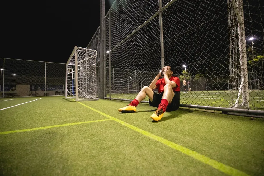 A footballer sitting against the fence after a lost match, head in hands